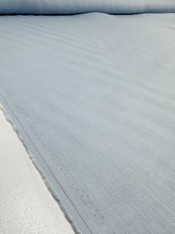 Close-up of Super Cheap Fabrics' Pure Linen in Powder Blue (140cm) on a white surface, showcasing its subtle weave pattern and red stitched edge. The lightweight, breathable material is neatly layered in smooth folds.
