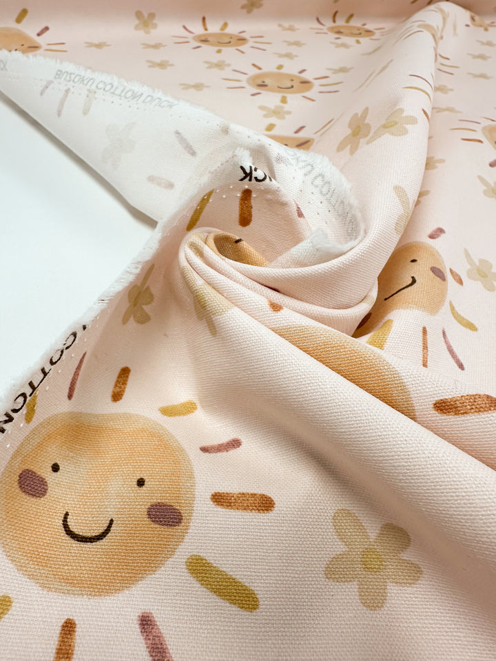 Close-up of Super Cheap Fabrics' Cotton Duck - Sunny - 112cm, featuring smiling sun faces, flowers, and abstract lines in warm pastels. Perfect for home décor projects with its soft texture; visible edge features printed text.