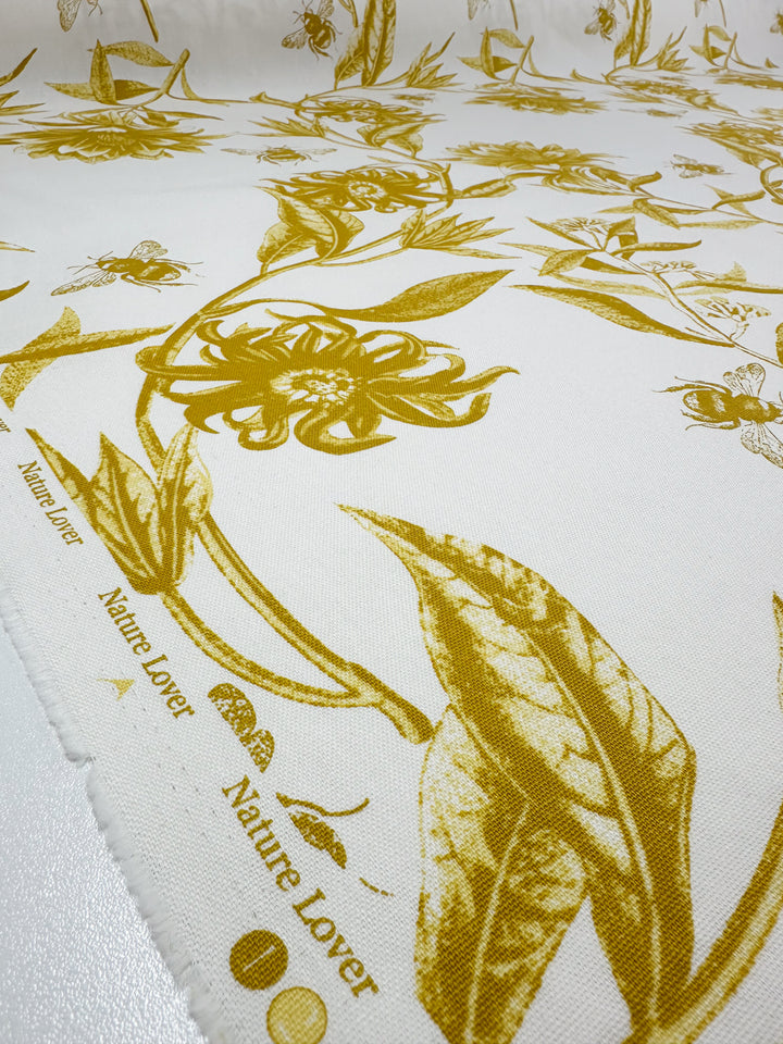 The Super Cheap Fabrics Cotton Duck - Bee Mustard is a charming textile featuring a golden floral and botanical print on white with "Nature Lover" elegantly repeated. Ideal for home décor, its detailed flowers, leaves, and insects bring charm to any space. Width: 150cm.