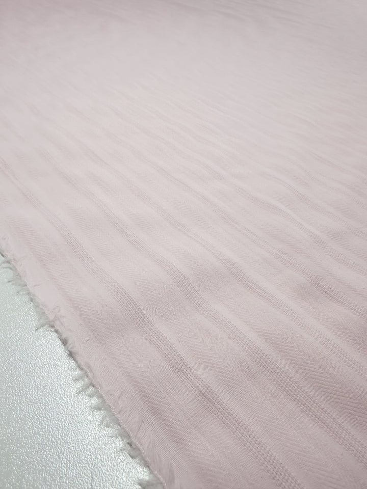 A Super Cheap Fabrics close-up: Textured Cotton - Silver Pink - 143cm. This fabric is 100% cotton with a wavy striped design on a smooth, pale surface, featuring slightly frayed edges.