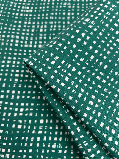 A close-up image of Stretch Bengaline - Green Glass fabric from Super Cheap Fabrics, featuring a white, grid-like pattern with small rectangular and square shapes. The medium-weight material is folded over itself, highlighting the texture and design of the 150cm wide fabric.