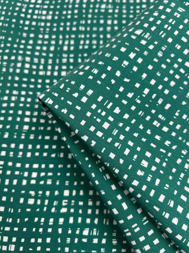 A close-up image of Stretch Bengaline - Green Glass fabric from Super Cheap Fabrics, featuring a white, grid-like pattern with small rectangular and square shapes. The medium-weight material is folded over itself, highlighting the texture and design of the 150cm wide fabric.