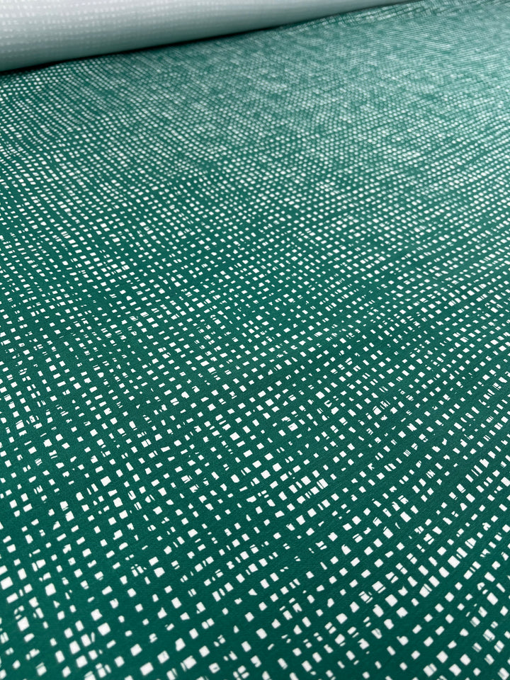 A close-up of Super Cheap Fabrics' "Stretch Bengaline - Green Glass - 150cm" shows a roll of green, medium-weight fabric adorned with a repeated small white square pattern. The scattered arrangement of the white squares creates a textured visual effect. The stretch bengaline fabric is unrolled and laid flat, occupying the majority of the frame.