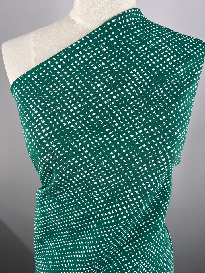 A mannequin adorned with Stretch Bengaline - Green Glass - 150cm from Super Cheap Fabrics, a medium-weight fabric featuring a white square grid pattern. The fabric is draped diagonally over the shoulder and around the body, showcasing its texture and design. The plain gray background accentuates the patterned material.
