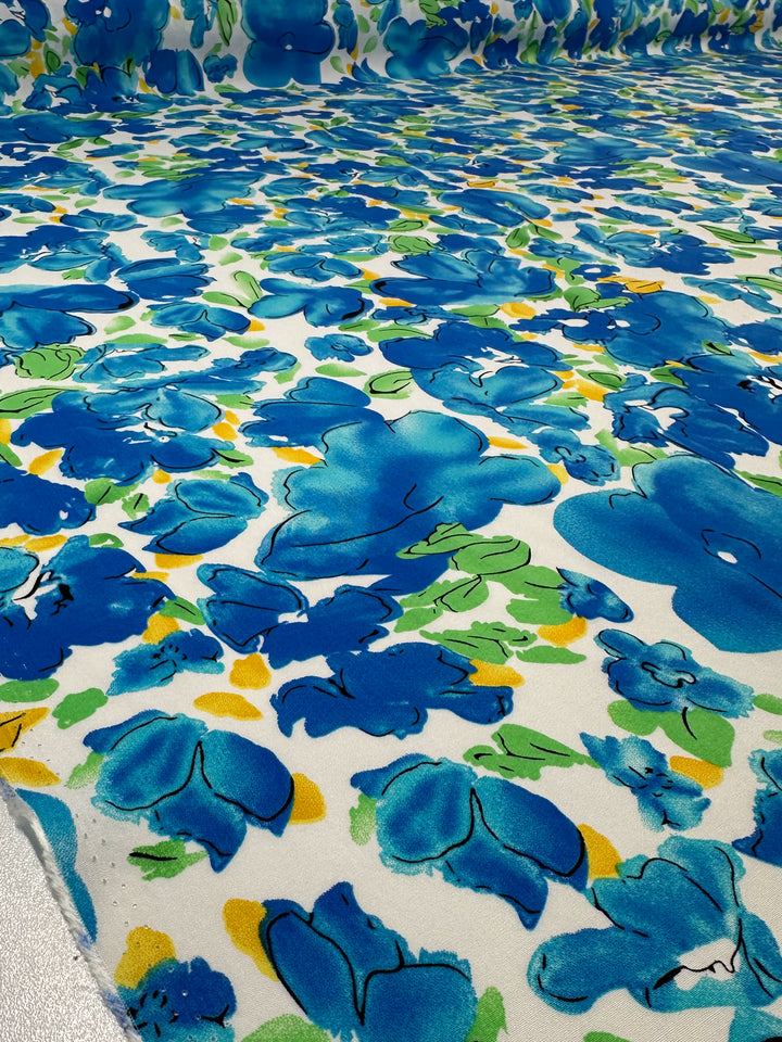 A close-up of Super Cheap Fabrics’ Designer Rayon - Azure Whispers (145cm), displaying vibrant blue abstract flowers and green-yellow leaves on a white background, with the striking print extending into the distance.