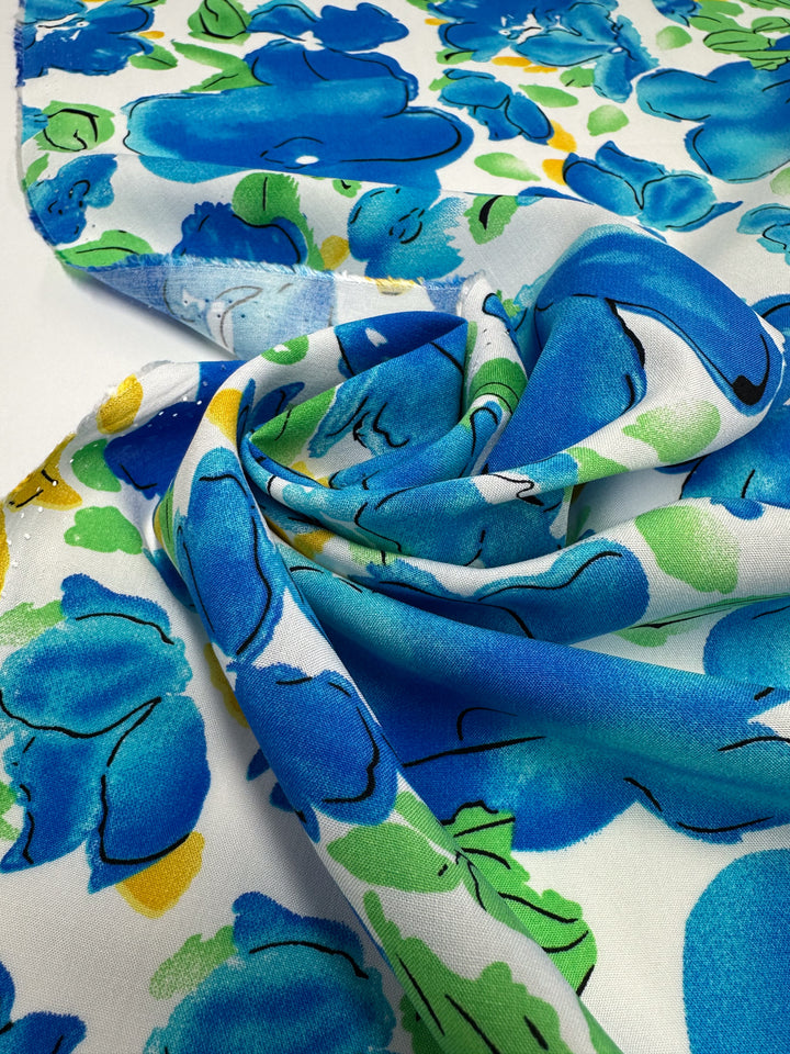 A close-up of Super Cheap Fabrics’ Designer Rayon - Azure Whispers (145cm) shows soft white fabric with bold, colorful floral prints—large blue flowers, green leaves, and small yellow accents—loosely gathered in gentle folds.