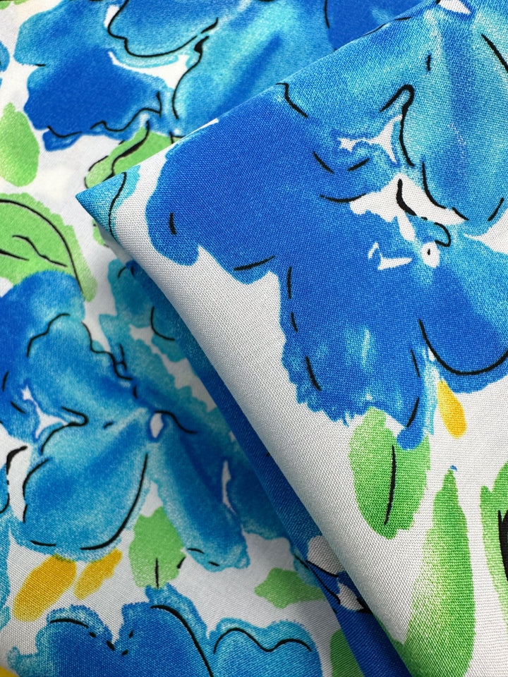 Close-up of Super Cheap Fabrics' Designer Rayon - Azure Whispers (145cm), showing its vivid blue floral print with green leaves and yellow accents on white. A folded section reveals the vibrant design from different angles.