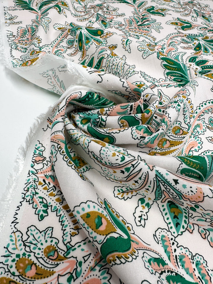 A close-up of Super Cheap Fabrics’ Designer Rayon - Verdant Script (145cm), featuring lightweight rayon with a vibrant green, pink, and yellow paisley floral pattern, slightly wrinkled and draped, and frayed edges on one side.