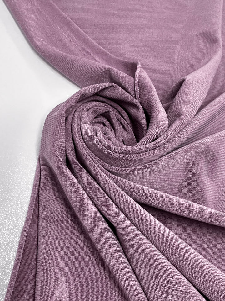 A close-up of Super Cheap Fabrics’ ITY Knit - Damson (150cm) shows soft folds and spiral swirls of light mauve polyester spandex, elegantly arranged on a smooth white surface.