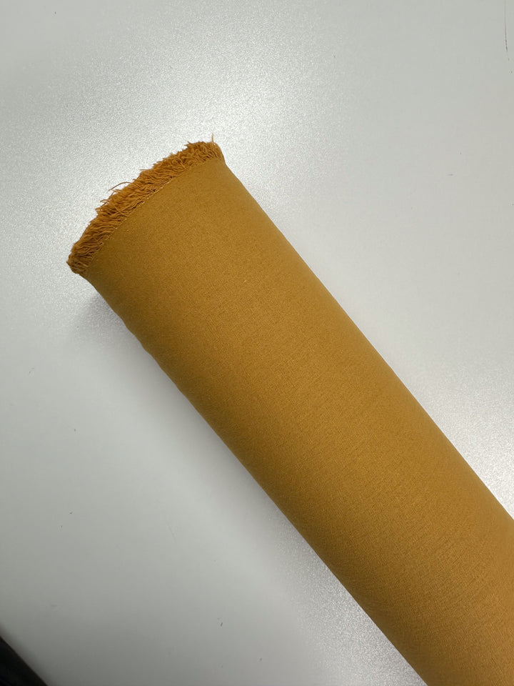 A roll of Ramie Linen in Inca Gold by Super Cheap Fabrics, measuring 150cm and featuring a frayed edge, lies on a smooth white surface.