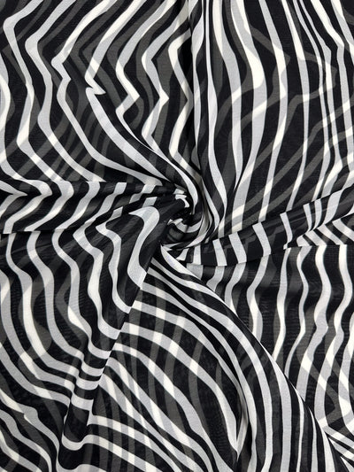The "Zebraic" Printed Chiffon by Super Cheap Fabrics features an abstract black-and-white pattern with swirling, wavy lines that create an optical illusion reminiscent of zebra stripes. This design on lightweight polyester adds a dynamic, three-dimensional feel, perfect for high fashion statements. Width: 150cm.