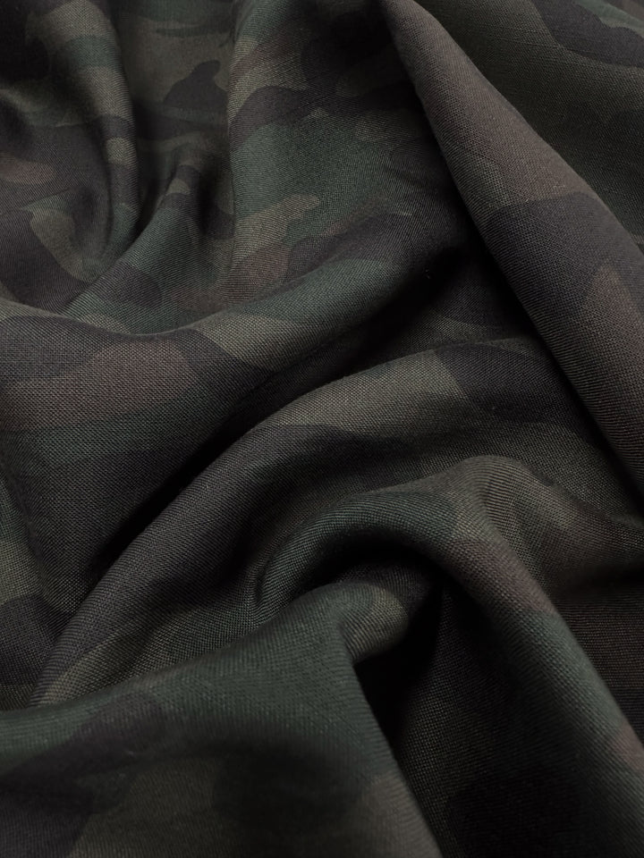 A close-up of Super Cheap Fabrics' Printed Linen - Military (150cm), featuring a dark green and black camouflage pattern, loosely draped to show gentle folds and shadows.