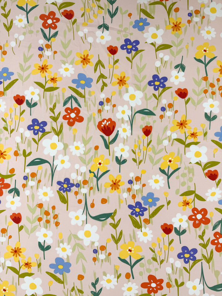 Designer Rayon - Meadow Mingle by Super Cheap Fabrics features a scattering of red, yellow, white, and blue wildflowers with green leaves on a soft pink background—perfectly versatile for any project.