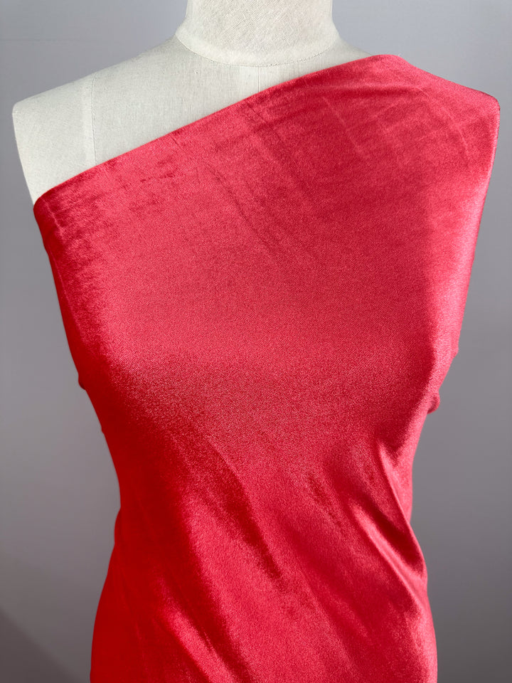 A mannequin showcases the vibrant "Rose of Sharon" Stretch Velour fabric by Super Cheap Fabrics. The glossy red, one-shoulder design flows smoothly, highlighting its sheen against a simple gray background.