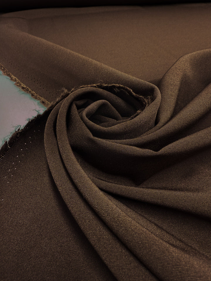 A close-up of Super Cheap Fabrics’ Cotton Bengaline - Mocha Earth - 150cm, medium weight with unfinished edges, shows its smooth texture and visible selvage as the stretch fabric lies flat, partially rolled and loosely gathered in a swirl.
