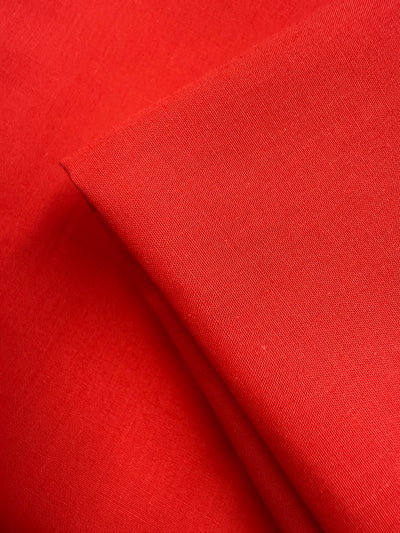 A close-up of two pieces of Super Cheap Fabrics' Poplin - Signal Red (114cm) shows smooth poly cotton texture and clean edges, with one piece slightly folded over the other.
