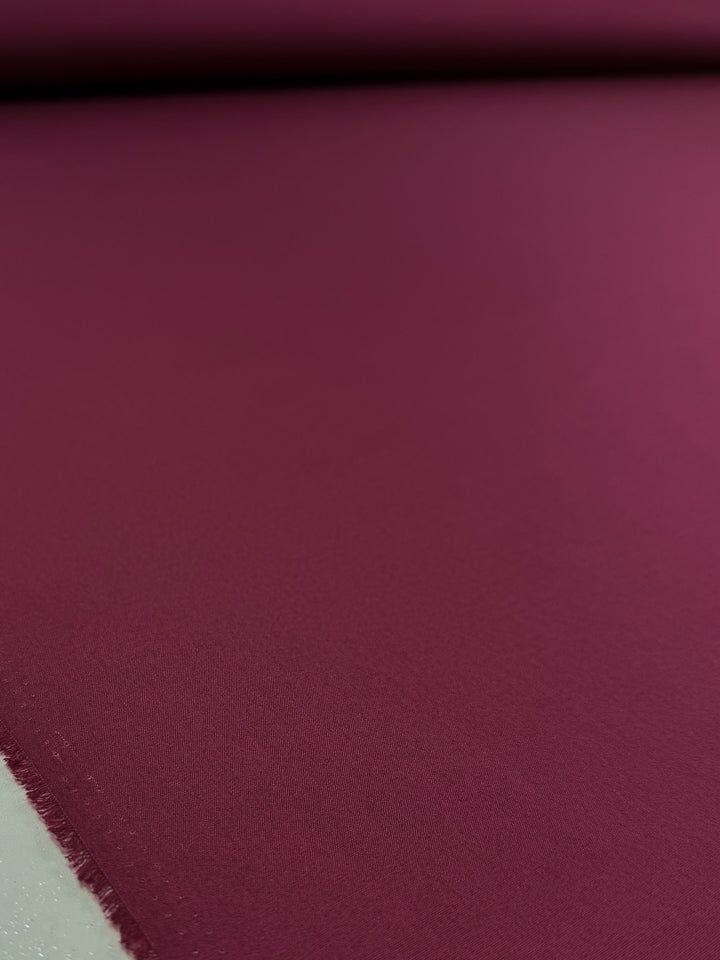 A close-up of Super Cheap Fabrics’ Poplin - Mulberry Matte - 114cm shows its smooth magenta fabric laid flat, featuring a slightly frayed edge along the left side.