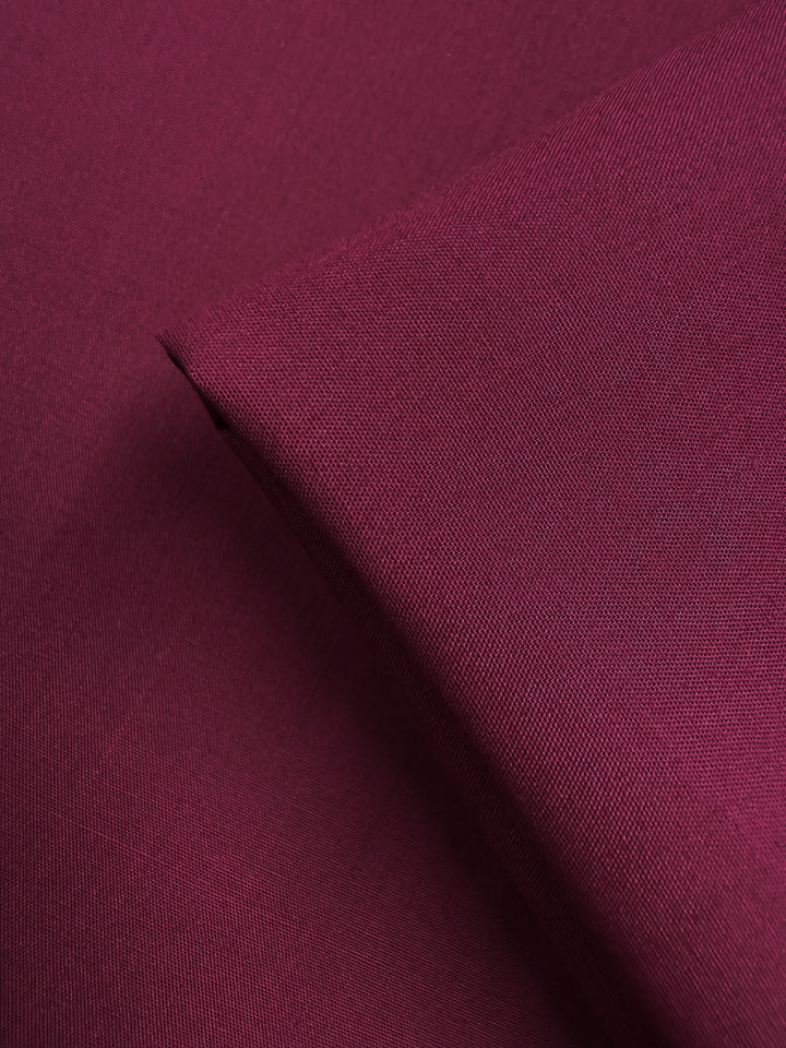 A close-up of Super Cheap Fabrics’ Poplin - Mulberry Matte (114cm) shows a rich maroon hue with visible weave texture; a folded corner highlights the smooth, matte finish and lightweight quality of this fabric.