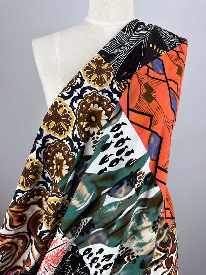 A mannequin draped in Super Cheap Fabrics' "Linen Blend - Patch - 145cm" showcases a colorful, patterned design. This breathable fabric features bold motifs including floral, abstract shapes, and geometric patterns in orange, brown, green, black, and white.