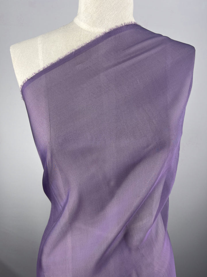 A mannequin is elegantly adorned with the extra lightweight, sheer "Silk Mousseline - Currant - 140cm" from Super Cheap Fabrics, wrapped diagonally from shoulder to waist. The fabric's delicate texture and soft lavender hue create a light, airy appearance against the plain gray background.