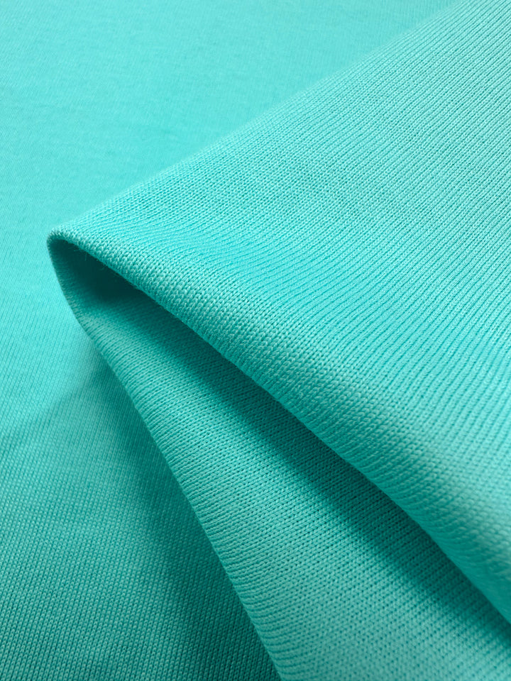 A close-up of Super Cheap Fabrics' Cotton Jersey in Aqua Splash shows its fine ribbed texture and vibrant hue. Its folds cast shadows highlighting the cotton's soft, smooth, and flexible nature.