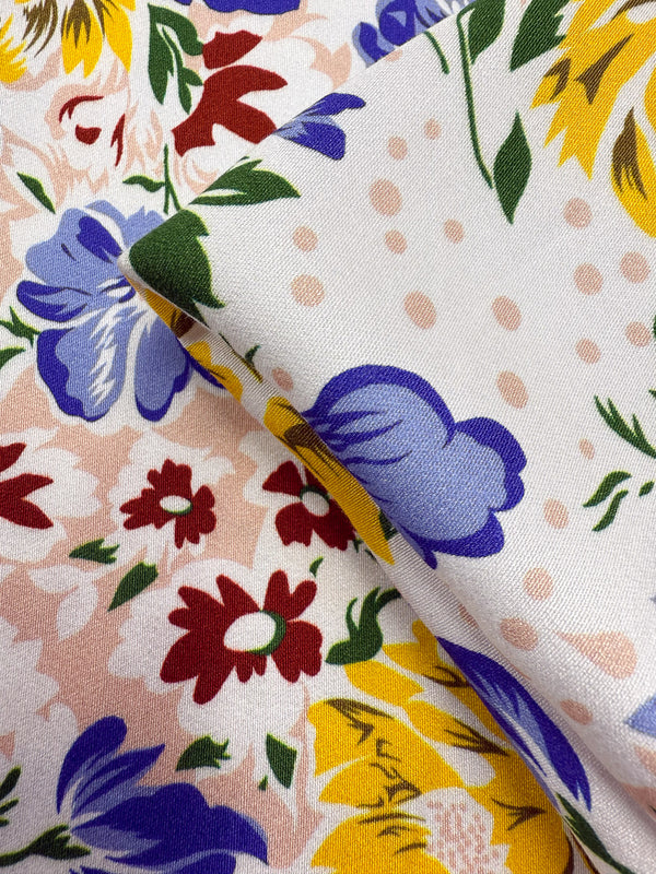 Close-up of Super Cheap Fabrics' Mercerised Rayon - Spring Bouquet (150cm): lightweight, breathable fabric with a multicolored floral print—red, yellow, and blue flowers, green leaves, and pink polka dots. Pattern shown on folded fabric.