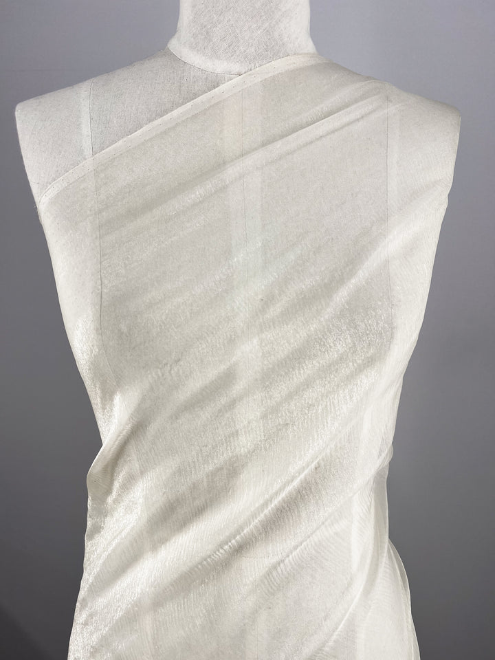 Super Cheap Fabrics' Silk Mousseline in Milk, measuring 137cm, elegantly drapes over a mannequin in an off-shoulder, asymmetrical style. Its extra-light weight and subtle texture are enhanced by a metallic shimmer against a neutral gray background.