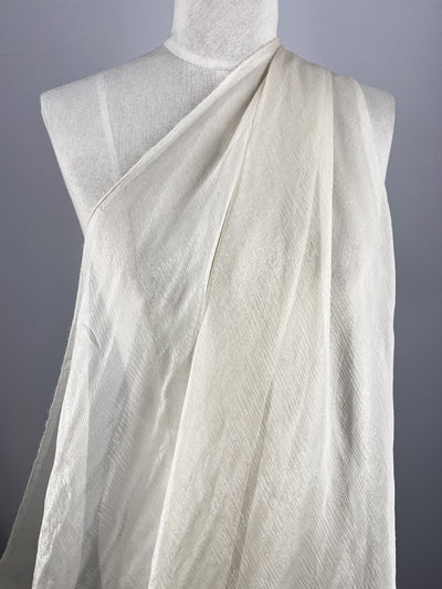 A white mannequin showcases Super Cheap Fabrics' Silk Mousseline in Milk, lightly draped over one shoulder for a flowing look with a subtle metallic shimmer against a plain gray backdrop.