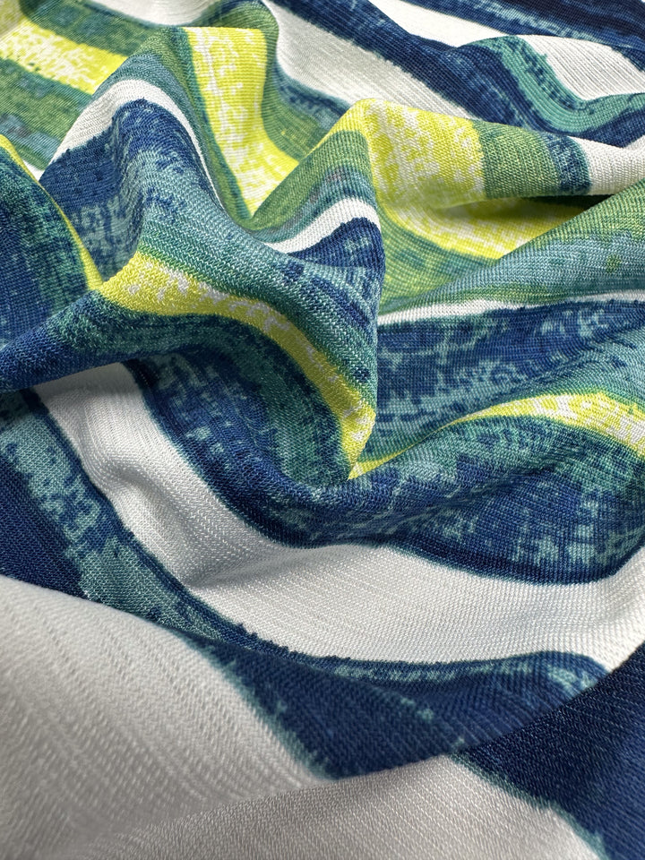 Close-up of Super Cheap Fabrics' Printed Lycra - Ocean Stripe - 145cm, showcasing a textured, wavy pattern with horizontal stripes in blue, white, green, and yellow. Its soft sheen and harmonious colors are ideal for dynamic summer garments.
