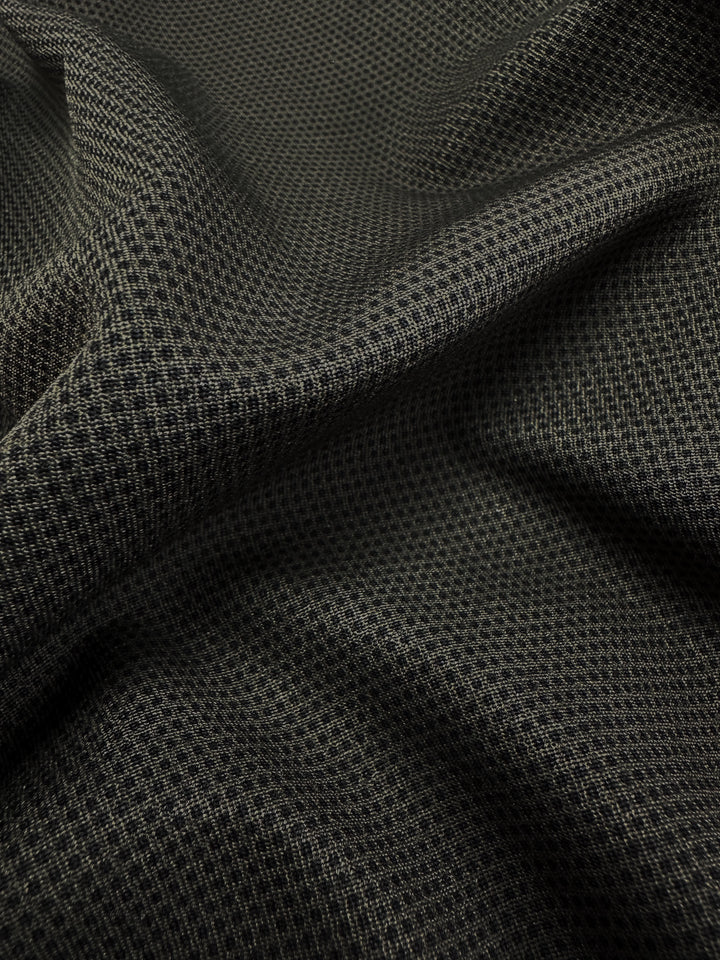 Close-up of Super Cheap Fabrics' Printed Bengaline - Pin Dot Khaki, a 150cm wide dark textured, medium-weight fabric with a small checkered pattern. The slightly wrinkled, stretchy material casts subtle shadows and highlights across its structured surface.