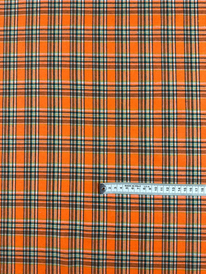 A piece of versatile fabric named "Suiting - Orange Glen Check - 147cm" from Super Cheap Fabrics features an orange, black, white, and green plaid pattern. A metallic measuring tape is laid across the lightweight fabric for scale, showing measurements up to 15 centimeters.