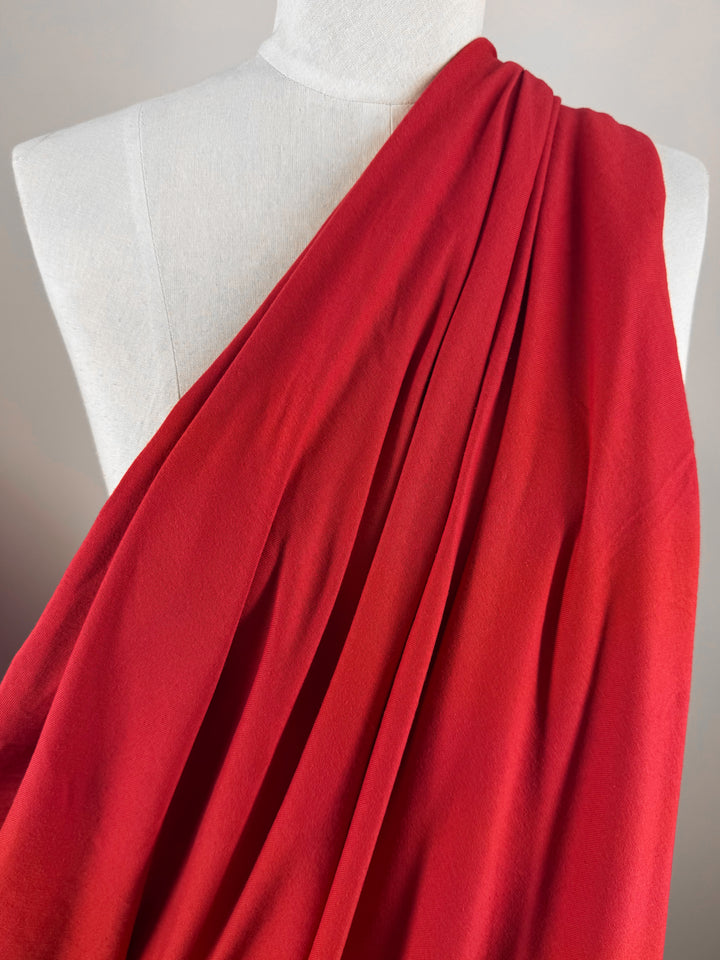 A close-up of a red cotton fabric named Cotton Jersey - Goji Berry - 165cm by Super Cheap Fabrics draped over a white mannequin, highlighting its flowing texture and vibrant goji berry color. It elegantly covers one shoulder, creating an asymmetrical look.