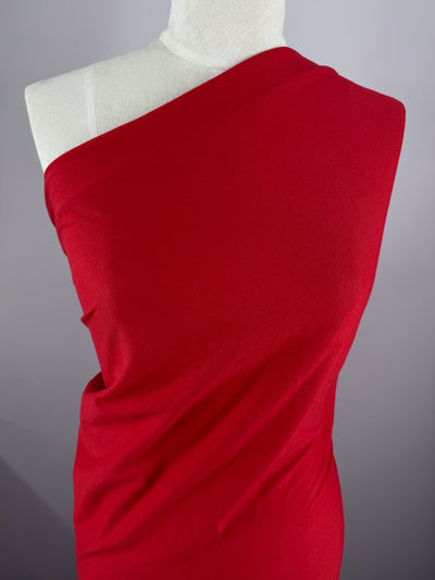 A mannequin showcases an elegant red, one-shoulder draped dress crafted from Super Cheap Fabrics' Cotton Jersey in a vibrant Goji Berry color. The smooth fabric flows gracefully from the right shoulder against a neutral gray backdrop, highlighting its striking hue.