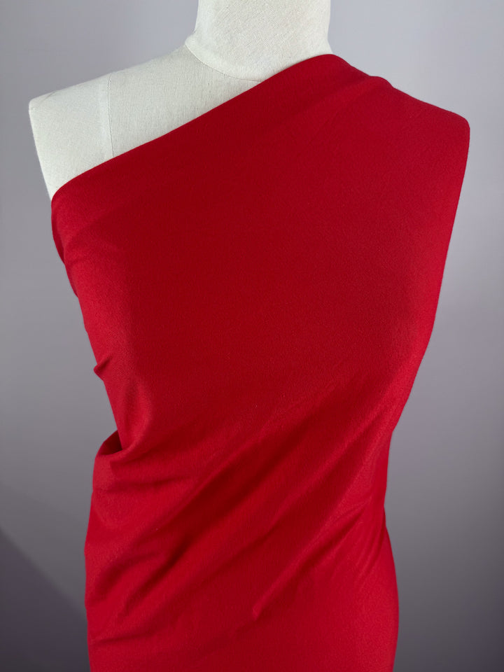 A mannequin showcases an elegant red, one-shoulder draped dress crafted from Super Cheap Fabrics' Cotton Jersey in a vibrant Goji Berry color. The smooth fabric flows gracefully from the right shoulder against a neutral gray backdrop, highlighting its striking hue.
