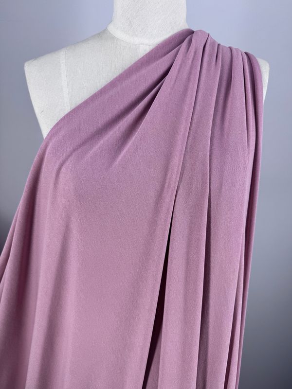 A dress form is adorned with Super Cheap Fabrics' Cotton Lycra in Fox Glove, draping elegantly over one shoulder. The soft, dusty pink material flows gracefully, revealing gentle folds and a smooth texture against a simple gray backdrop.