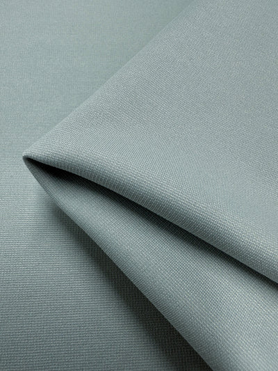 Close-up of neatly folded Ponte - Barely Blue fabric by Super Cheap Fabrics, measuring 155cm. The light gray material has a subtle texture and appears soft and stretchy, with gentle folds forming a triangular shape, highlighting its wrinkle-resistant quality against a plain background.