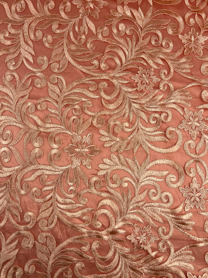 A close-up image of intricate, gold floral embroidery on a sheer coral lace fabric. The elaborate design features swirling patterns and blossoming flowers, reminiscent of Evening Lace - Galadriel from Super Cheap Fabrics, showcasing detailed craftsmanship and elegant, ornamental styling.