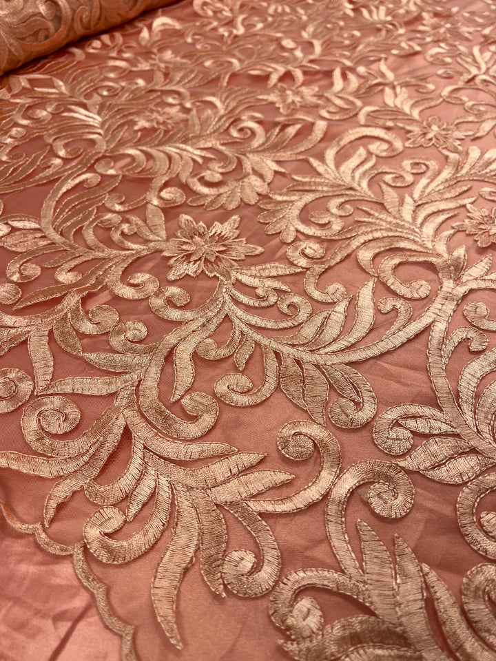 Close-up of an intricate, floral-patterned lace fabric in a soft peach color with shiny, golden embroidery. The design features swirling leaves, vines, and flowers, giving the Evening Lace - Galadriel by Super Cheap Fabrics an elegant and luxurious appearance.