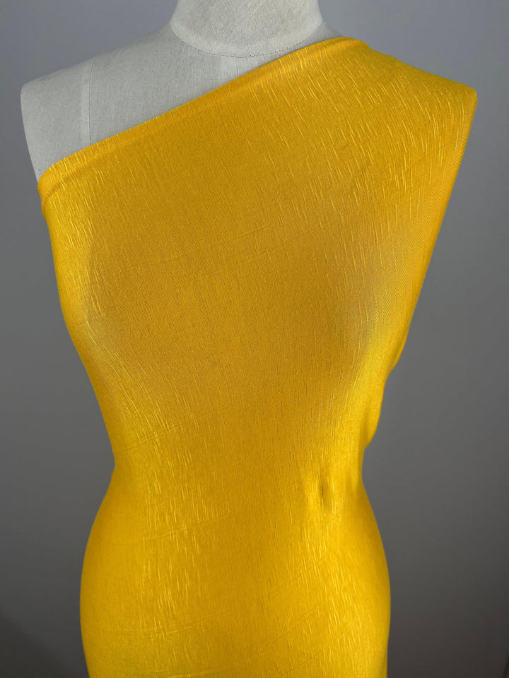 A mannequin is adorned with a vivid yellow one-shoulder dress made from Super Cheap Fabrics' Bamboo Jersey - Gold Fusion - 165cm. The material appears textured, adding subtle detail to the smooth and form-fitting design. The background is plain and grey, ensuring the focus remains entirely on this environmentally responsible dress.