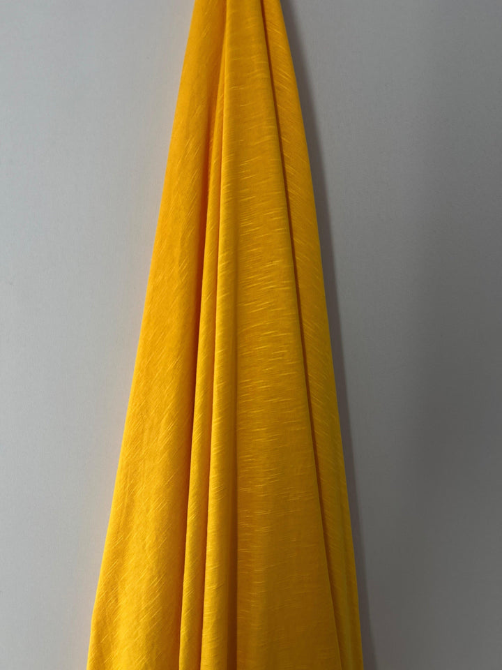 A bright yellow Bamboo Jersey - Gold Fusion - 165cm by Super Cheap Fabrics, silky to the touch, is draped against a plain, light gray background. The fabric has a slightly textured surface and hangs in smooth, natural folds.