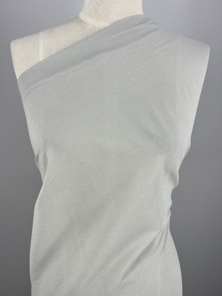 A dress form displays Super Cheap Fabrics' Stretch Crinkle - Mirage Gray - 140cm, a medium-weight polyester, draped one-shoulder style against a neutral gray background.
