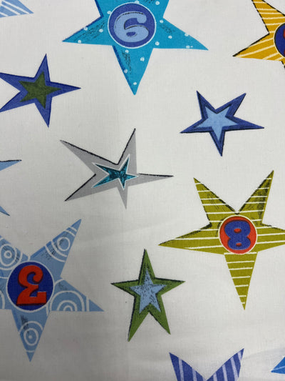 Super Cheap Fabrics' Printed Poplin - Number Star 112cm features a white poly-cotton base with blue, green, yellow, and gray stars. Each star contains a number—2, 3, 6, or 8—inside circles for a playful printed fabric design.