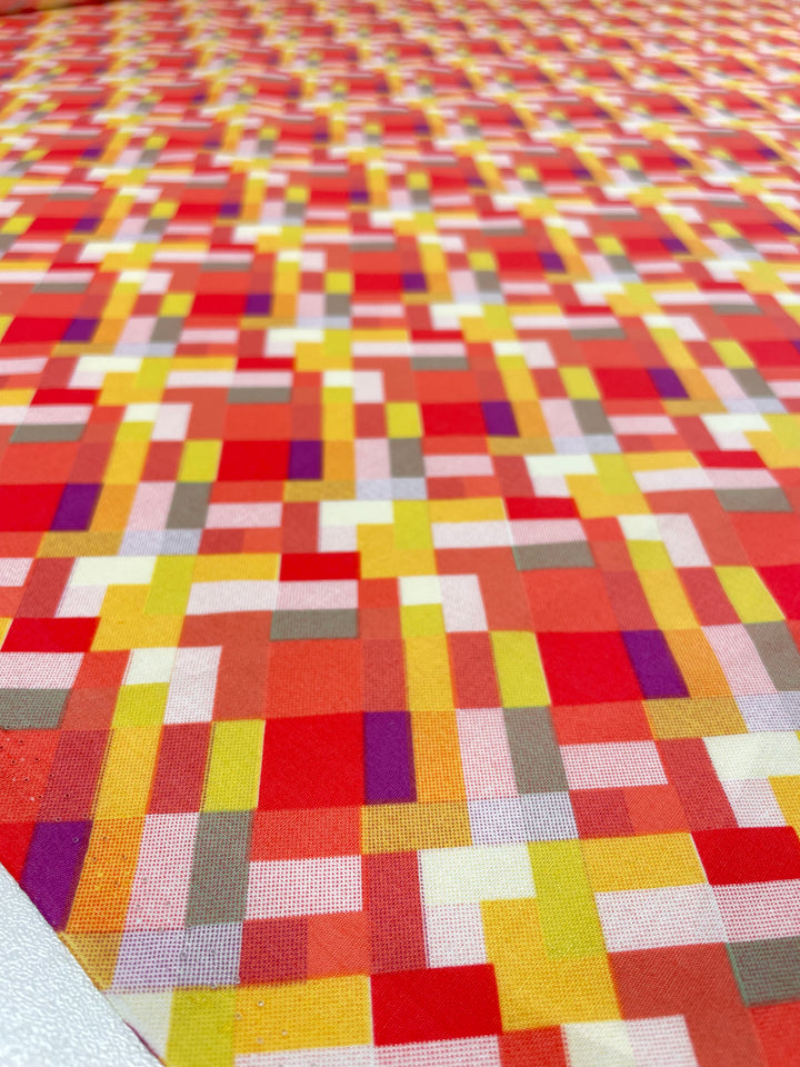The Printed Poplin - Jigsaw by Super Cheap Fabrics features a vibrant geometric checkered design in shades of red, orange, yellow, beige, and purple. This 112cm wide fabric is perfect for adding flair to any project or breathable clothing piece.